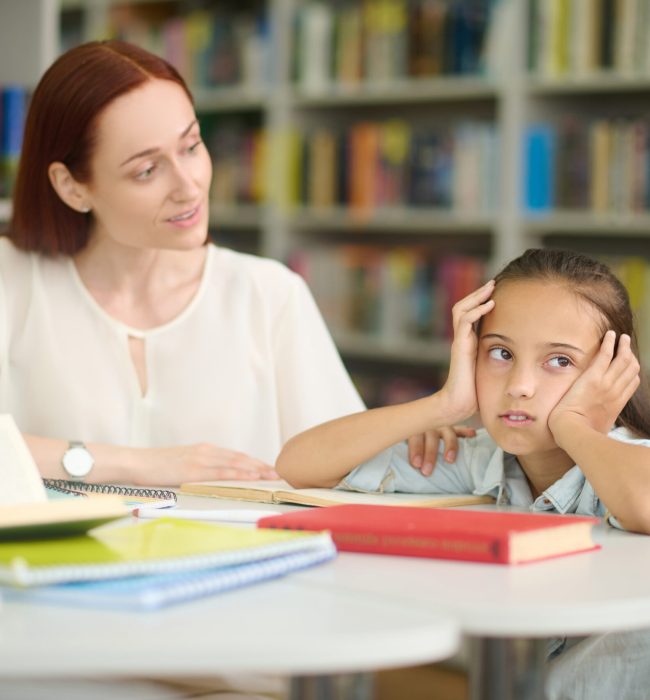 Overwork. Young caring woman looking questioningly at tired girl looking to side studying at table in room with bookshelves during day